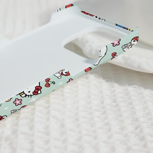 Puede incluir: Una funda de tel&eacute;fono blanca con un borde verde menta que presenta un patr&oacute;n repetido de personajes de dibujos animados, cerezas y flores. La funda tiene un acabado liso y mate y est&aacute; dise&ntilde;ada para un tel&eacute;fono inteligente.