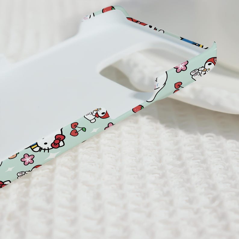 Puede incluir: Una funda de tel&eacute;fono blanca con un borde verde menta que presenta un patr&oacute;n repetido de personajes de dibujos animados, cerezas y flores. La funda tiene un acabado liso y mate y est&aacute; dise&ntilde;ada para un tel&eacute;fono inteligente.