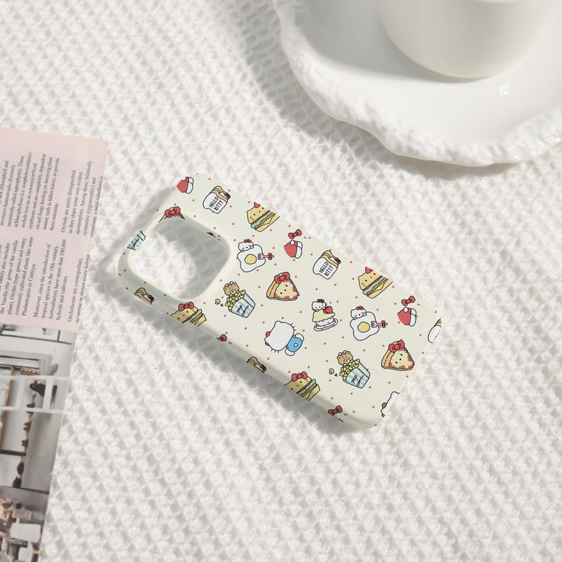 May include: A light-colored phone case featuring a repeating pattern of Hello Kitty characters, food items, and red bows. The case has a cutout for the camera and buttons. The background is a white, textured surface.