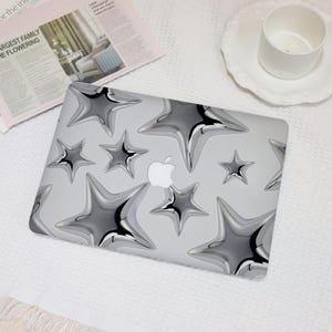 May include: A silver laptop case with a pattern of metallic star shapes. The stars have a three-dimensional appearance and are arranged across a light grey background. The Apple logo is visible in the centre.