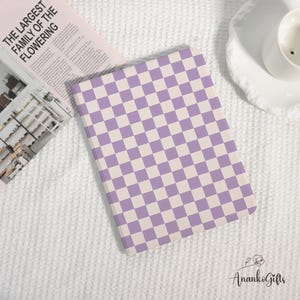 May include: A notebook featuring a purple and white chequered design. The notebook is placed on a white textured surface, alongside a magazine with the text "THE LARGEST FAMILY OF THE FLOWERING". The notebook has a soft cover.