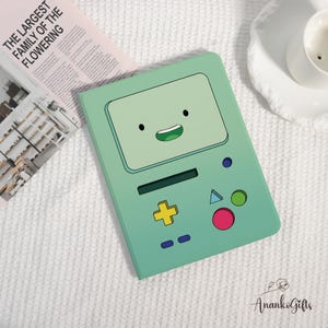 May include: A mint green tablet case featuring a cartoon face with a smiling mouth and black eyes. The case has a yellow cross, a blue triangle, a green circle, and a pink circle. The text "THE LARGEST FAMILY OF THE FLOWERING" is visible on a pink newspaper.