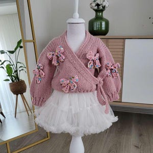 May include: A pink knitted wrap sweater with bow embellishments and a white tulle skirt. The sweater features a wrap design and is adorned with multiple decorative bows. The skirt is layered and fluffy, creating a classic look.
