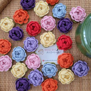 Könnte beinhalten: Eine Sammlung farbenfroher, gehäkelter Blumenapplikationen in verschiedenen Farbtönen wie Orange, Lila, Rot, Gelb, Rosa und Blau. Jede Blume hat eine grüne Basis und ist auf einer gewebten Oberfläche angeordnet. Eine Karte mit dem Text "Rosemoon Knit Boutique" ist ebenfalls vorhanden.