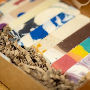 Könnte beinhalten: Eine Schachtel gefüllt mit bunten, rechteckigen Seifenstücken. Die Seifen haben verschiedene Designs und Farben, darunter blaue, weiße, braune und Regenbogenmuster. Braune Papierschnitzel umgeben die Seifen, und die Schachtel ist aus Pappe.