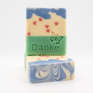 Könnte beinhalten: Zwei handgemachte Seifen, eine mit einem blauen und weißen Wirbelmuster und die andere mit einem grünen und weißen Muster mit roten Herzen und dem Wort "Danke" in grün.