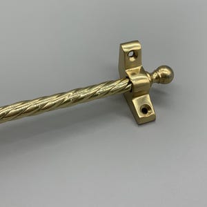 May include: A brass window stay featuring a decorative twisted rod and a ball finial. The stay has a rectangular base with two screw holes. The brass has a polished finish.