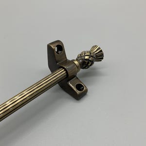 May include: A brass curtain pole with a fluted design and a thistle finial. The pole is mounted to a wall bracket.