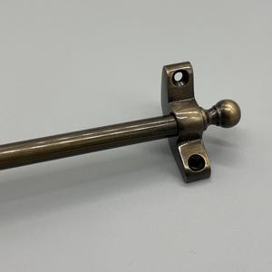 May include: Close-up of an antique brass window stay. The stay features a long, cylindrical rod connected to a mounting bracket with two screw holes. A decorative ball adorns the end of the rod, adding a touch of elegance.