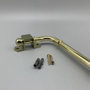 May include: A gold metal curtain rod with a rounded end and a bracket. The rod is bent at a 90-degree angle. The bracket has a screw hole and is attached to the rod with a screw. The rod is 30 cm long. The bracket is 5 cm wide. The rod is 1.25 cm in diameter. The rod is made of metal and has a gold finish. The rod is used to hang curtains.