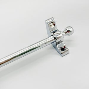 May include: A polished chrome window stay with a ball finial. The stay has a rectangular base with two screw holes and a cylindrical rod. The ball finial is on the end of the rod. The window stay is designed to hold a window open.