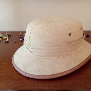 Op de afbeelding: Een beige safarihoed met een brede rand en een bruine leren rand. De hoed heeft een gestructureerde kroon met een band en oogjes. De hoed ligt op een houten oppervlak.