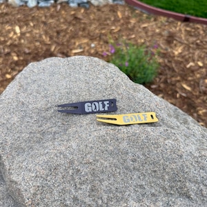 Op de afbeelding: Twee golfdivot-reparatietools. Eén is donkerblauw met het woord "GOLF" in wit, en de andere is geel met dezelfde tekst. Beide tools liggen op een grote, grijze rots.