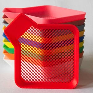 May include: A collection of colourful paint trays in shades like red, pink, brown, grey, blue, yellow, green, and orange. The front tray is red and features a grid. These trays are designed for holding paint.