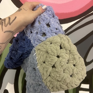 Op de afbeelding: Een handgemaakte gehaakte deken in blauw- en groentinten. De deken is gemaakt van zacht, dik garen en heeft een granny square design. De vierkanten zijn aan elkaar gestikt met witte draad. De deken wordt tegen een kleurrijke achtergrond gehouden.