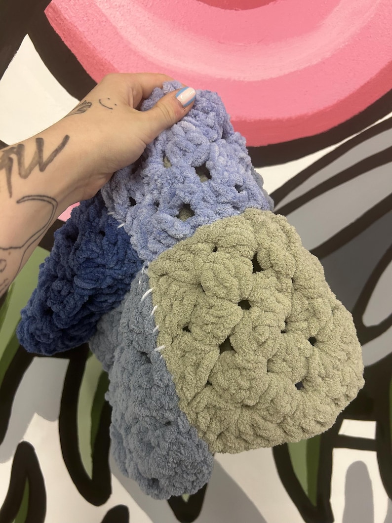 Op de afbeelding: Een handgemaakte gehaakte deken in blauw- en groentinten. De deken is gemaakt van zacht, dik garen en heeft een granny square design. De vierkanten zijn aan elkaar gestikt met witte draad. De deken wordt tegen een kleurrijke achtergrond gehouden.