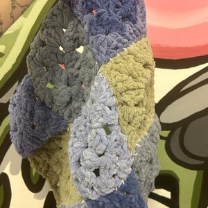 Op de afbeelding: Een handgemaakte, dikke gehaakte deken in een patchwork-ontwerp. De deken heeft vierkanten in verschillende tinten blauw, grijs en olijfgroen. Het garen heeft een zachte, pluche textuur en de steken zijn zichtbaar.