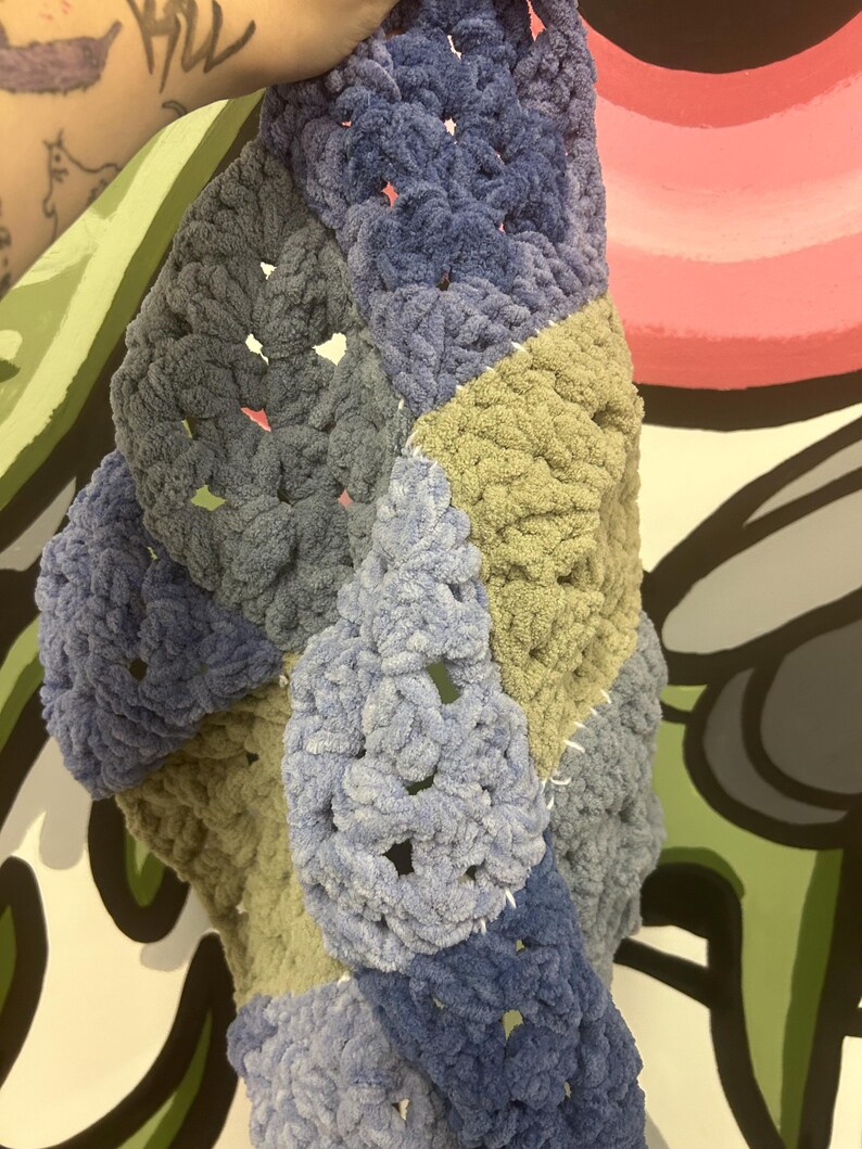 Op de afbeelding: Een handgemaakte, dikke gehaakte deken in een patchwork-ontwerp. De deken heeft vierkanten in verschillende tinten blauw, grijs en olijfgroen. Het garen heeft een zachte, pluche textuur en de steken zijn zichtbaar.