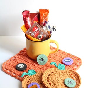 May include: A yellow mug filled with coffee packets and candy sits on an orange crocheted placemat. Decorative wooden Halloween-themed coasters with spider, skull, and pumpkin designs are also present.