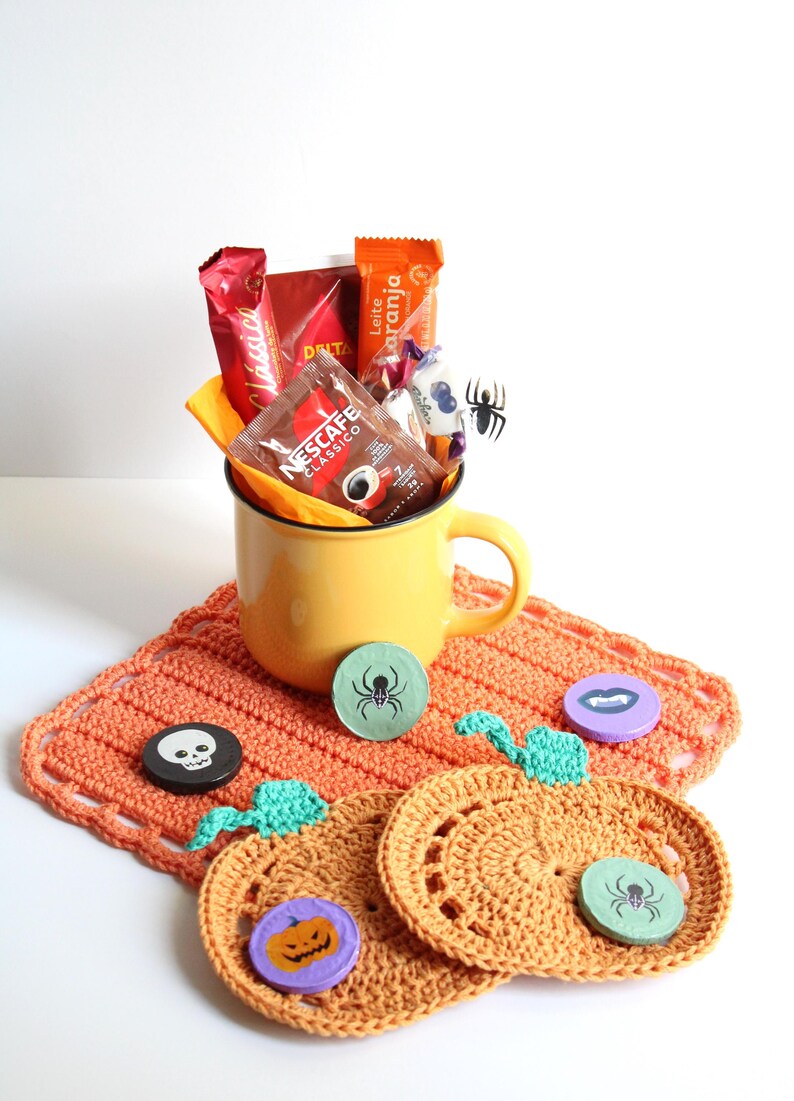May include: A yellow mug filled with coffee packets and candy sits on an orange crocheted placemat. Decorative wooden Halloween-themed coasters with spider, skull, and pumpkin designs are also present.