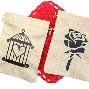 May include: Two beige burlap drawstring bags with black designs. One bag features a cage with a heart, the other a rose. A red crocheted item is between the bags. The bags are on a white background.
