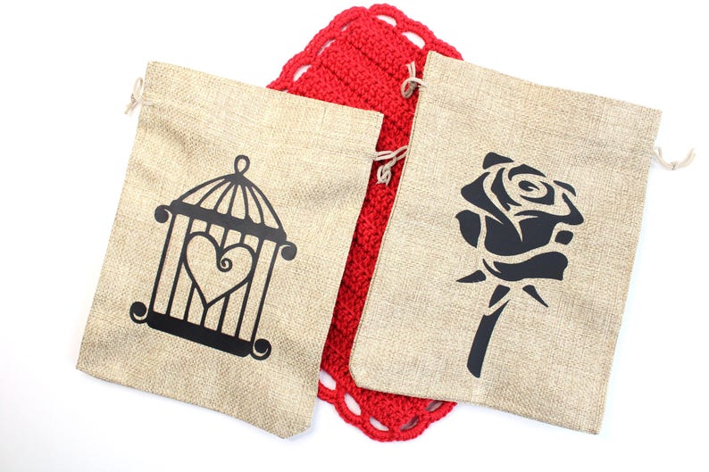 May include: Two beige burlap drawstring bags with black designs. One bag features a cage with a heart, the other a rose. A red crocheted item is between the bags. The bags are on a white background.