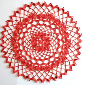 Può includere: Un centrino all'uncinetto rotondo di colore arancione vivace caratterizzato da un intricato motivo a reticolo traforato. Il design si irradia da un motivo centrale ispirato a un fiore, espandendosi verso l'esterno con forme geometriche.
