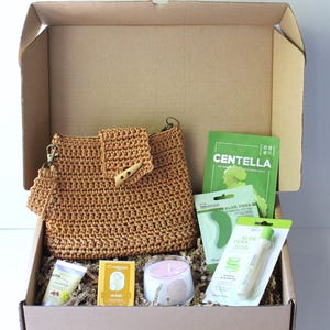 May include: A gift box containing self-care products. It features a brown crocheted handbag, skincare items, a candle, and face masks. The box is filled with shredded paper for cushioning.