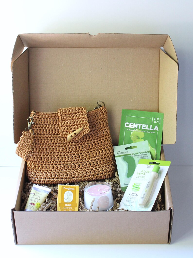 May include: A gift box containing self-care products. It features a brown crocheted handbag, skincare items, a candle, and face masks. The box is filled with shredded paper for cushioning.