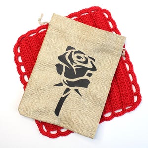 May include: A beige burlap drawstring bag with a black rose design, resting on a red crocheted coaster with a scalloped edge. The bag is centered, and the coaster is visible beneath it.