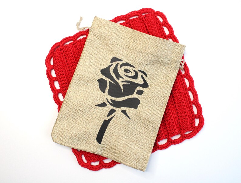 May include: A beige burlap drawstring bag with a black rose design, resting on a red crocheted coaster with a scalloped edge. The bag is centered, and the coaster is visible beneath it.