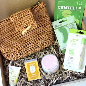 May include: A gift box containing beauty products. It features a woven brown handbag, skincare items like aloe vera eye pads and a roll-on eye serum, a face mask, and a small candle. The box is lined with shredded paper.