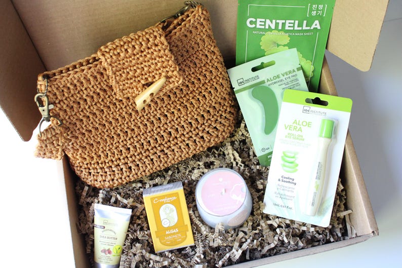 May include: A gift box containing beauty products. It features a woven brown handbag, skincare items like aloe vera eye pads and a roll-on eye serum, a face mask, and a small candle. The box is lined with shredded paper.