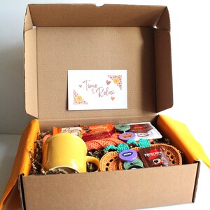 May include: A cardboard box filled with relaxation items. Inside, a yellow mug, crochet coasters, and a small card that reads "Time to Relax". The box also contains snacks and coffee packets.