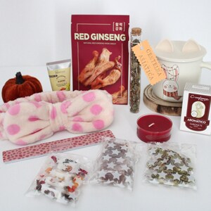 May include: A collection of self-care items, including a plush pink polka-dot headband, a red candle, a mug with a bunny design, and a package of Red Ginseng face mask. Other items include a nail file and small bags of decorative items.