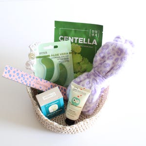 Pu&ograve; includere: Un cestino intrecciato pieno di prodotti di bellezza. Include una lima per unghie rosa e blu, patch per gli occhi, una maschera per il viso, un tubetto di crema e una fascia per la testa con stampa leopardata lavanda. I prodotti sono disposti su sfondo bianco.