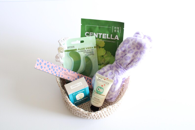 Pu&ograve; includere: Un cestino intrecciato pieno di prodotti di bellezza. Include una lima per unghie rosa e blu, patch per gli occhi, una maschera per il viso, un tubetto di crema e una fascia per la testa con stampa leopardata lavanda. I prodotti sono disposti su sfondo bianco.
