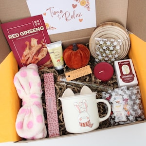 May include: A gift box filled with self-care items. Includes a red ginseng face mask, a small orange pumpkin, a white mug with a bunny design, a pink headband with polka dots, a red candle, and other items.