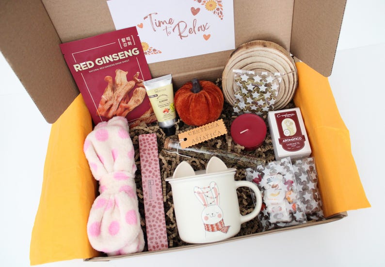 May include: A gift box filled with self-care items. Includes a red ginseng face mask, a small orange pumpkin, a white mug with a bunny design, a pink headband with polka dots, a red candle, and other items.