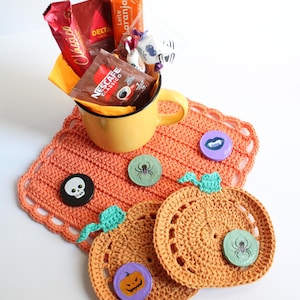 May include: A yellow mug filled with coffee packets and candy sits on an orange crocheted placemat. Two crocheted pumpkin coasters and decorative pins with Halloween designs are also present. A festive, handmade gift set.