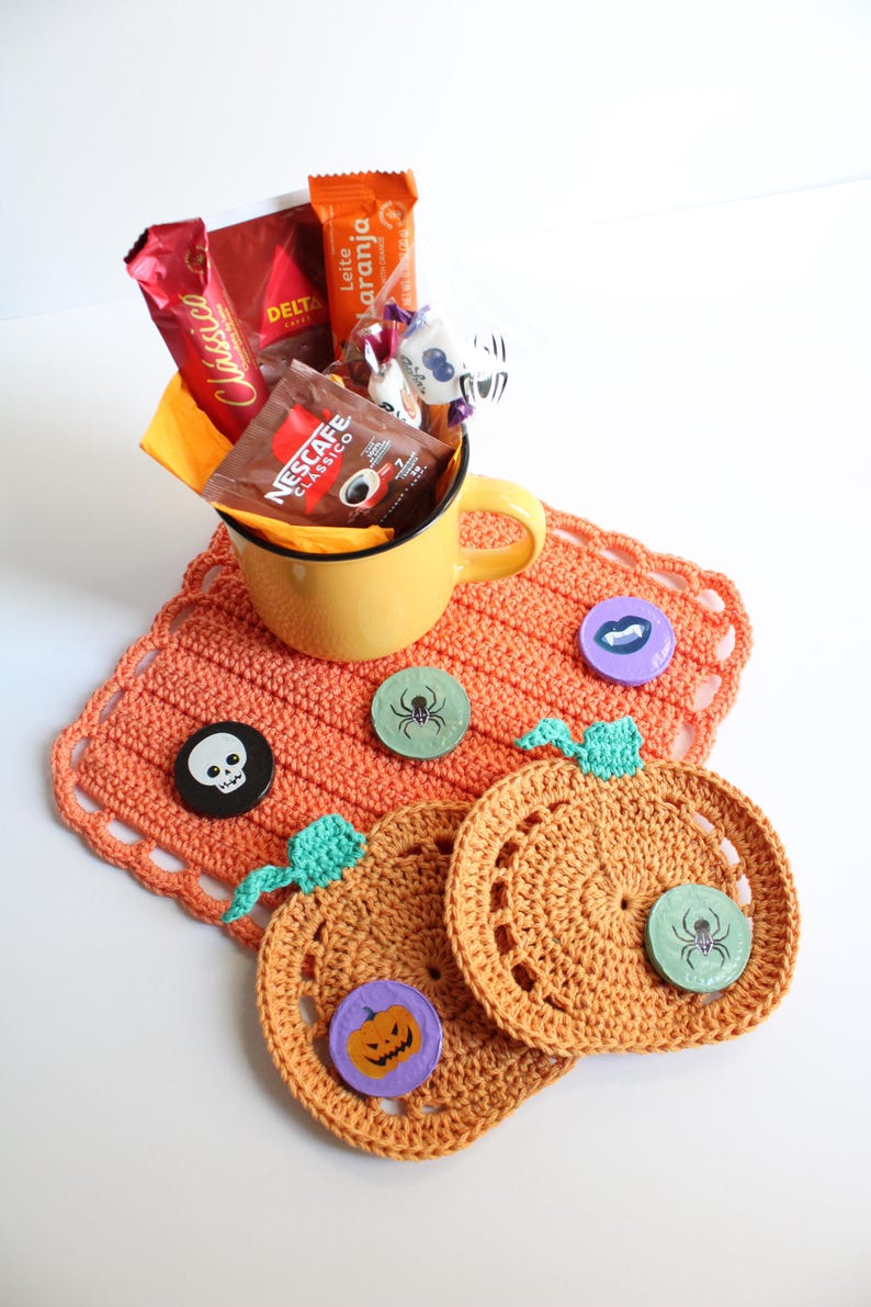 May include: A yellow mug filled with coffee packets and candy sits on an orange crocheted placemat. Two crocheted pumpkin coasters and decorative pins with Halloween designs are also present. A festive, handmade gift set.