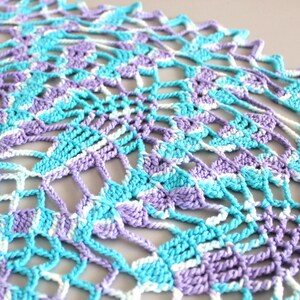 Pu&ograve; includere: Primo piano di un centrino all'uncinetto fatto a mano nei toni dell'azzurro, della lavanda e del bianco. L'intricato design traforato presenta un motivo geometrico ripetuto. Il centrino &egrave; realizzato con filato morbido.