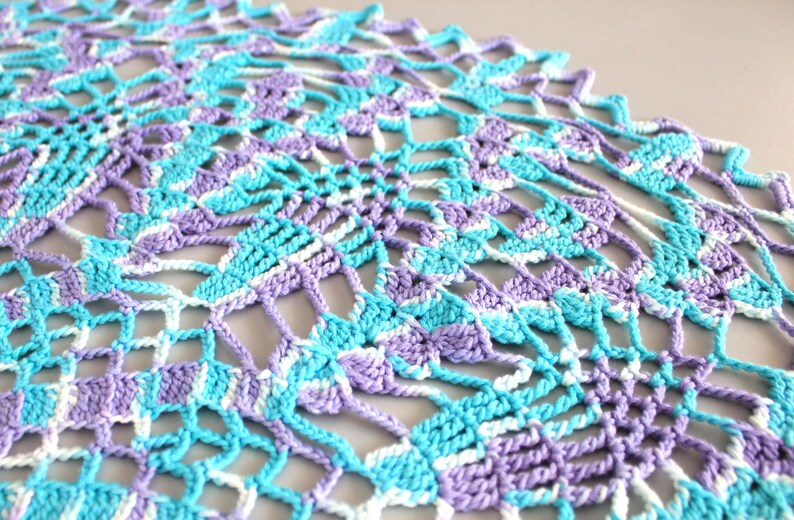 Pu&ograve; includere: Primo piano di un centrino all'uncinetto fatto a mano nei toni dell'azzurro, della lavanda e del bianco. L'intricato design traforato presenta un motivo geometrico ripetuto. Il centrino &egrave; realizzato con filato morbido.