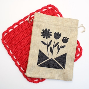 May include: A beige burlap drawstring bag with a black floral design, on a red crocheted coaster. The bag features a black envelope with flowers. The coaster has a scalloped edge.