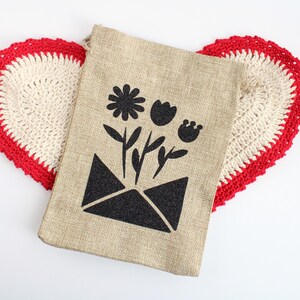 May include: A beige burlap bag featuring a black floral design, including a daisy, tulip, and bud, emerging from a black envelope. The bag rests on a heart-shaped, cream crocheted doily with a red border.
