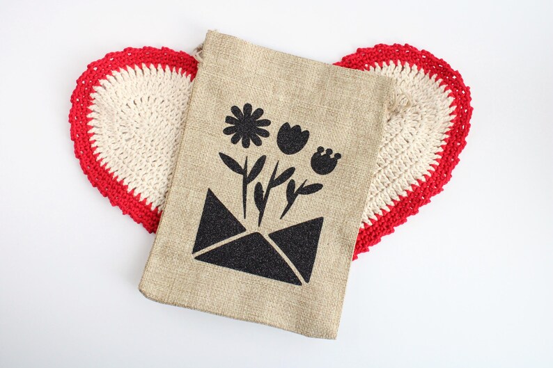May include: A beige burlap bag featuring a black floral design, including a daisy, tulip, and bud, emerging from a black envelope. The bag rests on a heart-shaped, cream crocheted doily with a red border.