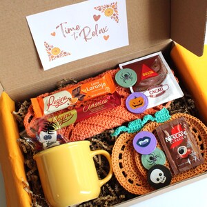 May include: A gift box filled with a yellow mug, chocolates, coffee, and decorative pins. The box contains a crocheted pumpkin-shaped coaster and a card that reads "Time to Relax". The box is lined with brown paper.