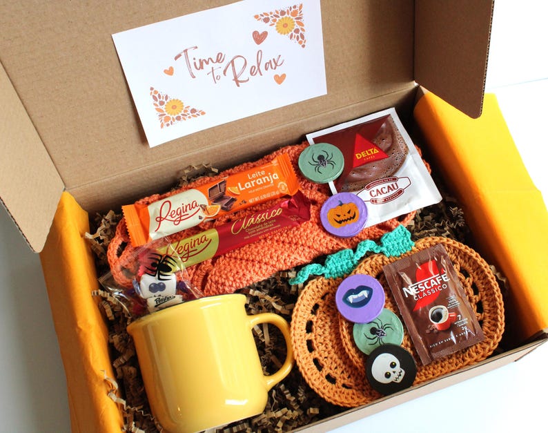 May include: A gift box filled with a yellow mug, chocolates, coffee, and decorative pins. The box contains a crocheted pumpkin-shaped coaster and a card that reads "Time to Relax". The box is lined with brown paper.