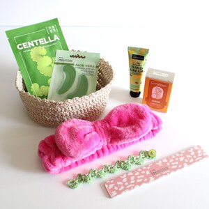 May include: A collection of beauty and self-care products, including a pink headband, a woven basket with face masks, hand cream, and a nail file. The face masks are labeled "Centella" and "Aloe Vera". The nail file has a pink and white pattern.