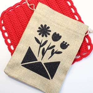 May include: A beige burlap drawstring bag featuring a black floral design, including a flower and leaves, over a geometric shape. The bag is placed on a red, crocheted, square doily with a scalloped edge.